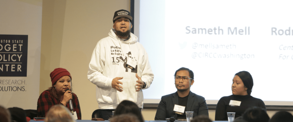 four people behind a desk, one man standing addressing attendees.