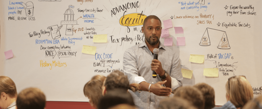 Man standing and speaking with microphone in front of audience, which art piece in background.